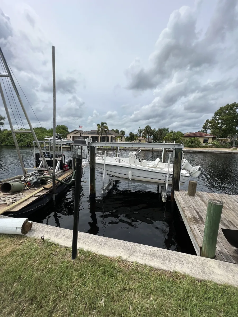 Boat lift and timber pilings beside a lawn and canal under a cloudy sky.
