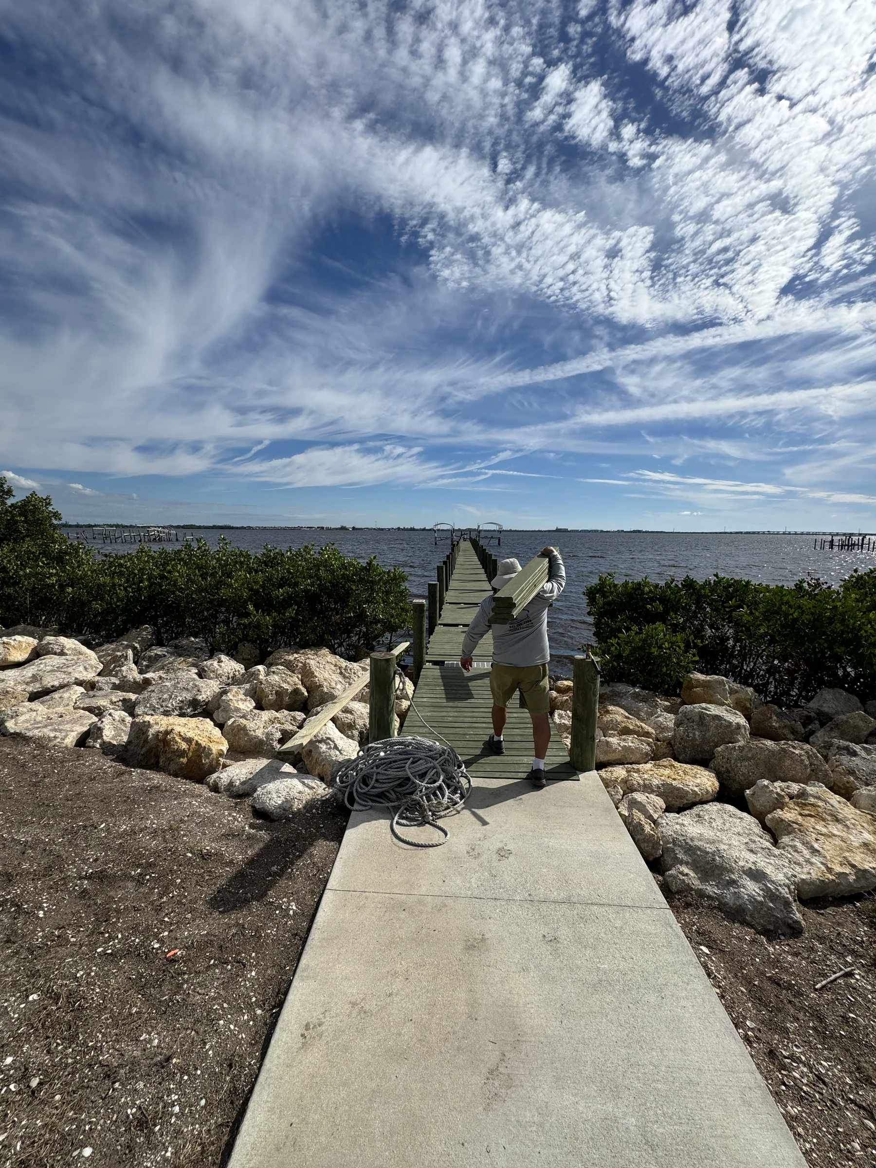 Crew member carrying boards toward a dock beside shoreline rocks and open water