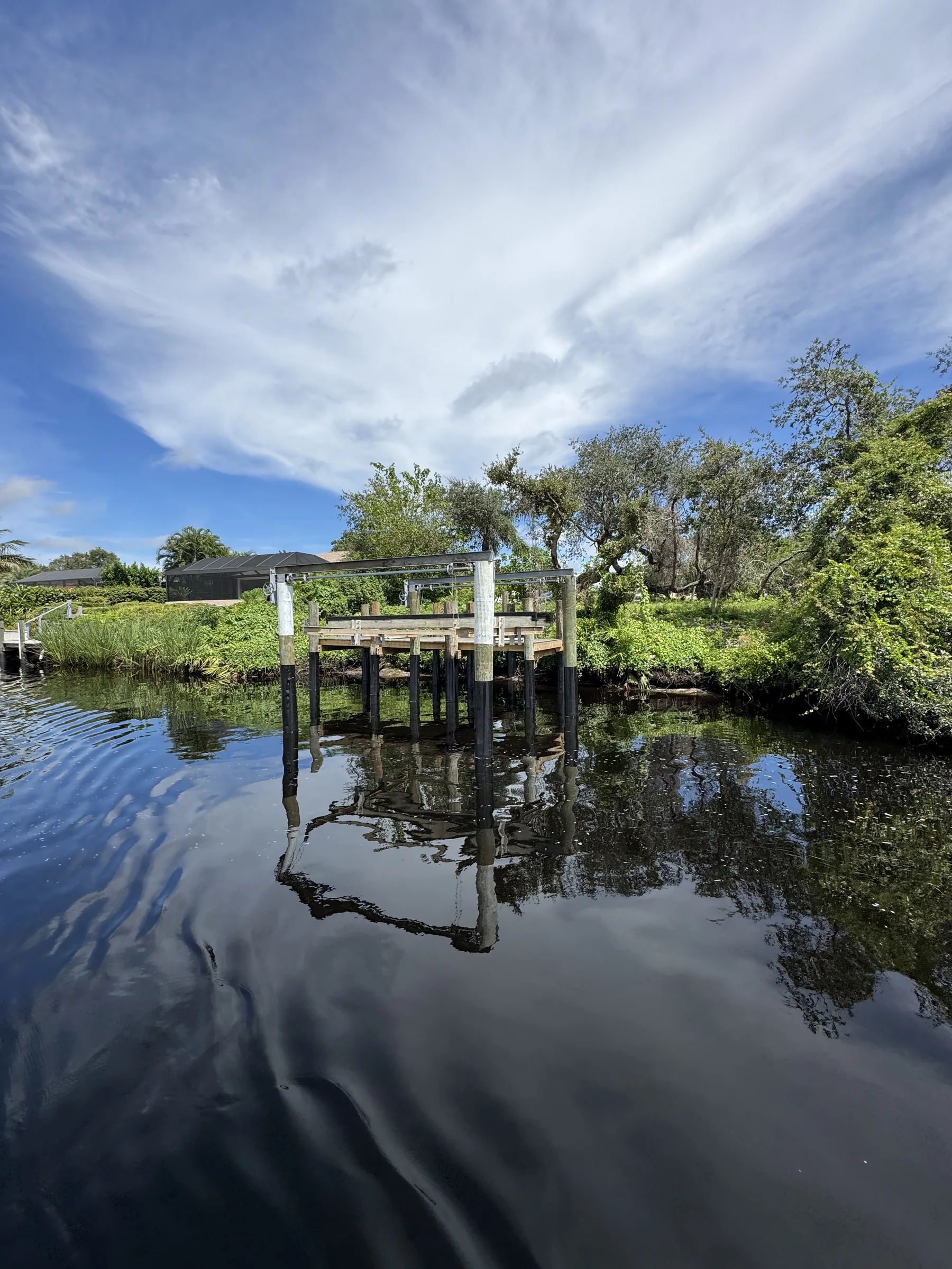 A boat lift reflected in calm water along a green residential shoreline
